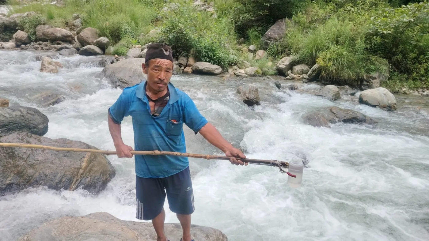 A field technician stands in fast-flowing water holding a long-handled bucket sampler to collect a river sediment sample