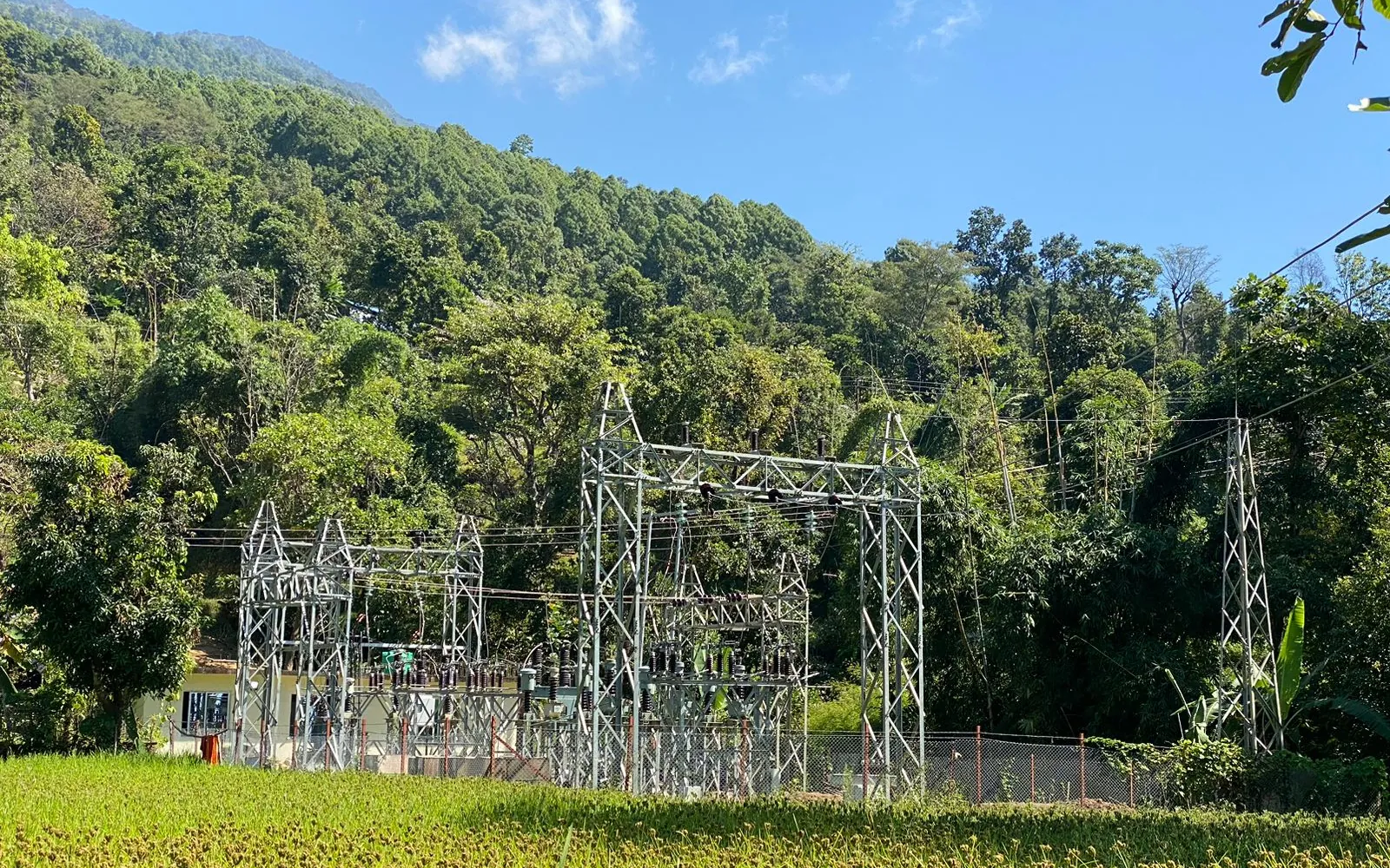 Switchyard structures and a transmission tower beside green fields in a forested valley at Machha Khola